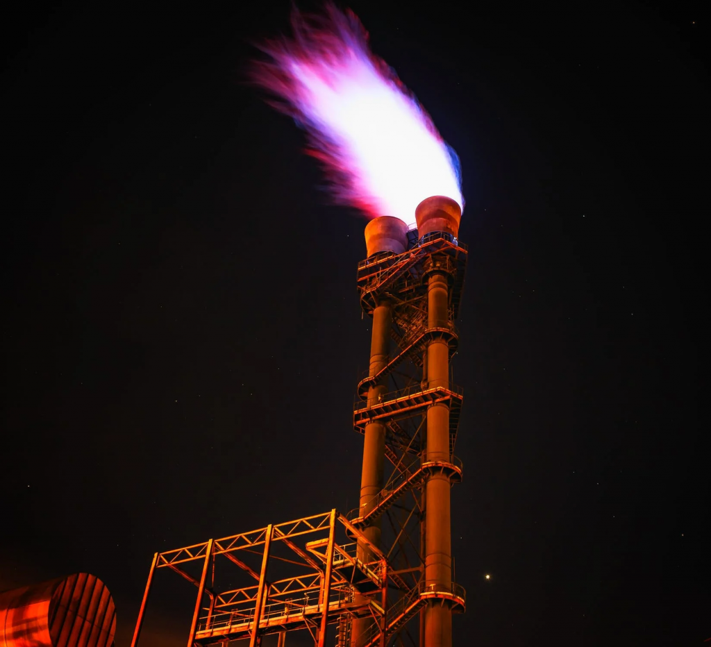 Vista de ángulo bajo de una torre de quema de gases tóxicos iluminada contra el cielo nocturno. Los gases provocan cancer en las poblaciones cercanas.